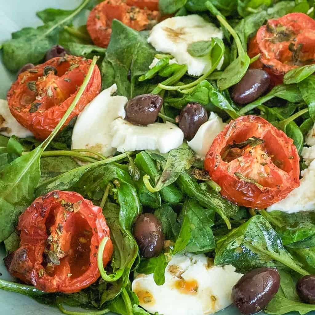 arugula spread on a blue platter, topped with roasted tomato halves, cheese and olives