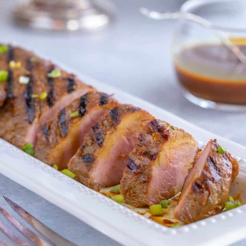 White platter filled with slices of Asian Pork Tenderloin. Bowl if rice and candles in the background