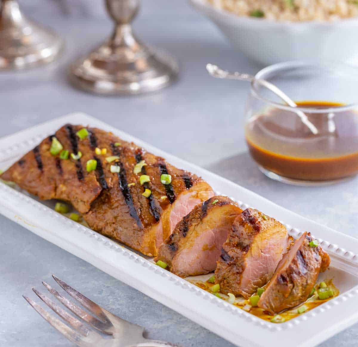 white platter filled with slices of Asian Pork Tenderloin. Small bowl of rice and candles in the background