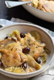 two braised chicken thighs with olives and artichoke hearts in a shallow bowl