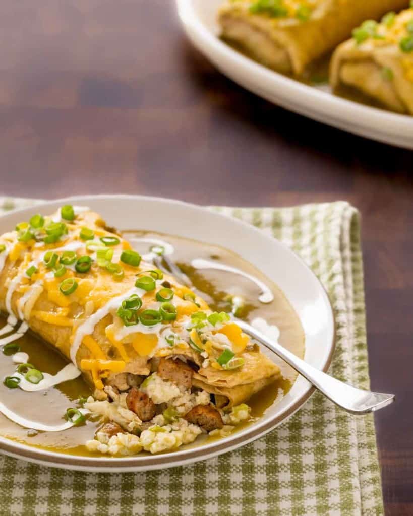 plate holding one breakfast burrito smothered in green chile sauce, with a corner open and two more on a platter in the backgrond
