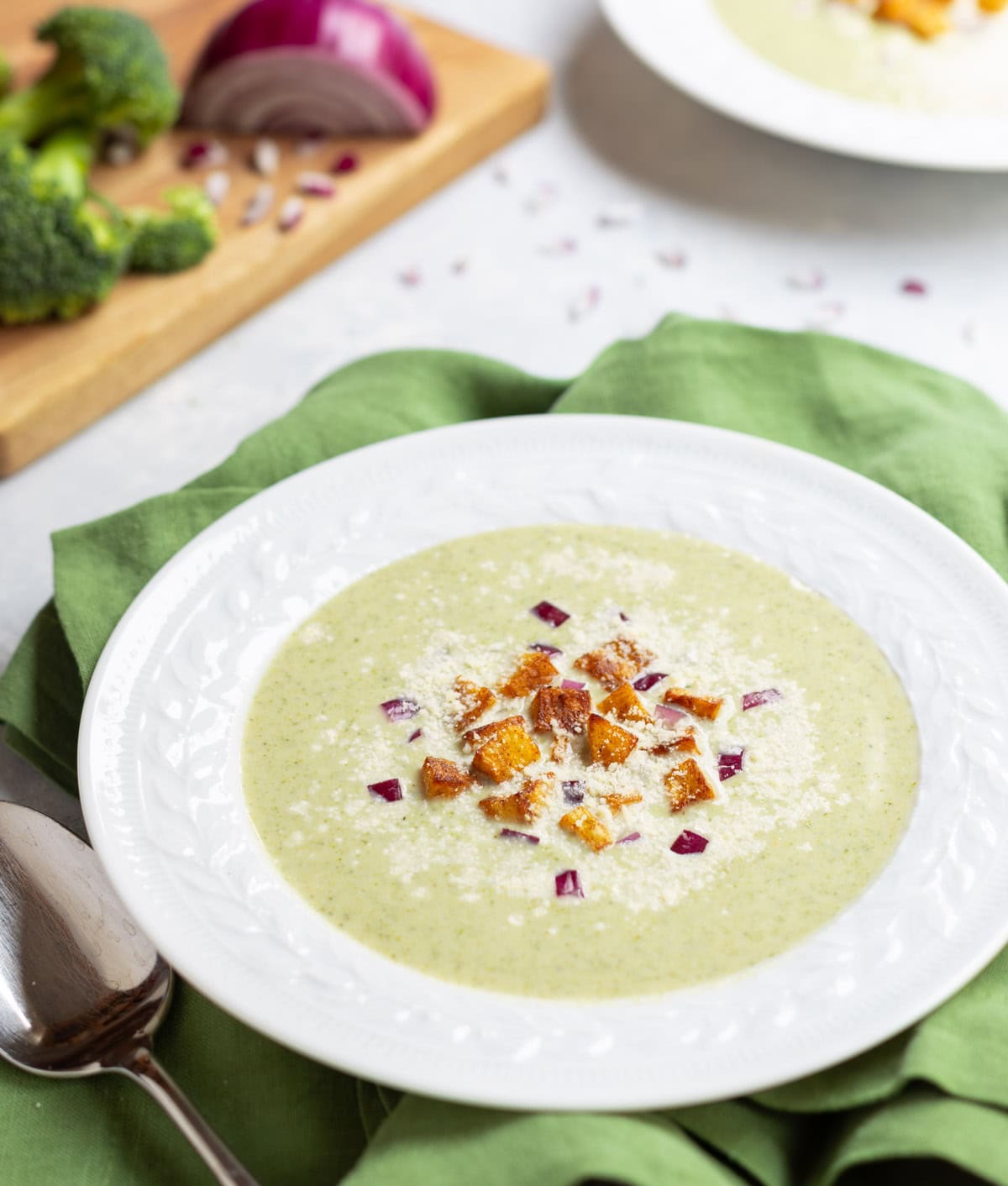 white bowl holding one serving of Healthy Broccoli Soup with potato croutons, red onion and Parmesan cheese garnishes. broccoli florets and partially chopped red onion in the background, along with a second bowl of soup