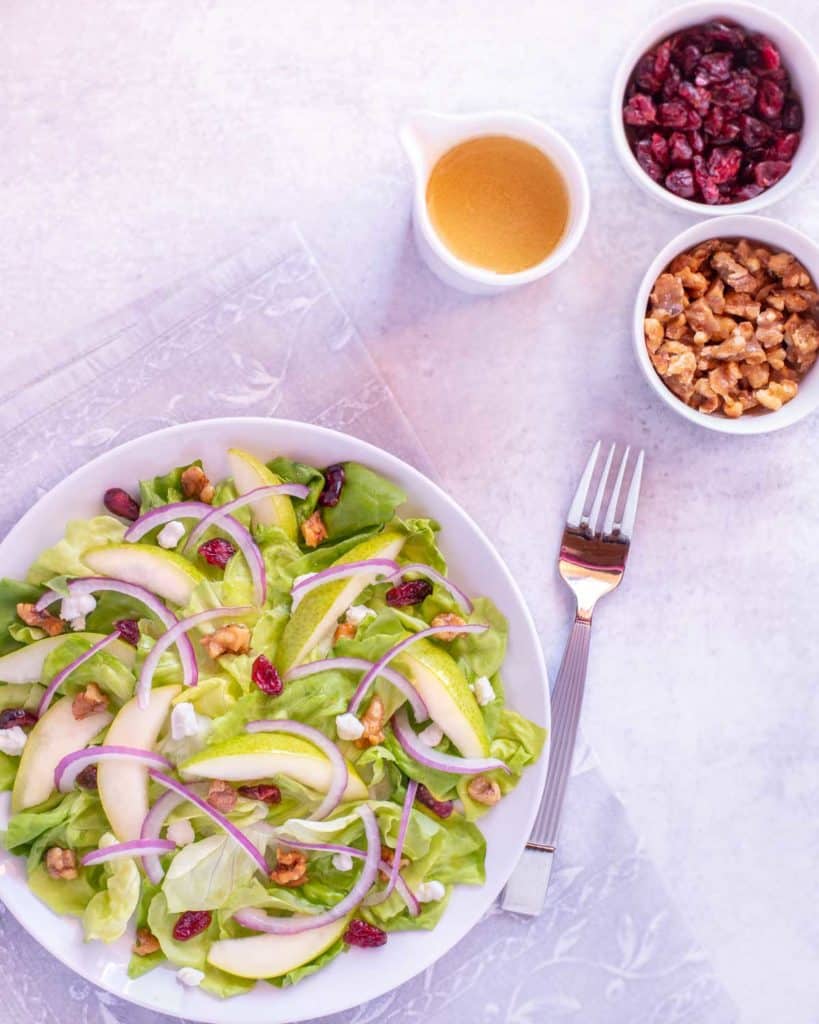 Plate showing Pear and Goat Cheese Salad, with dressing, candied walnuts and dried cranberries on the side