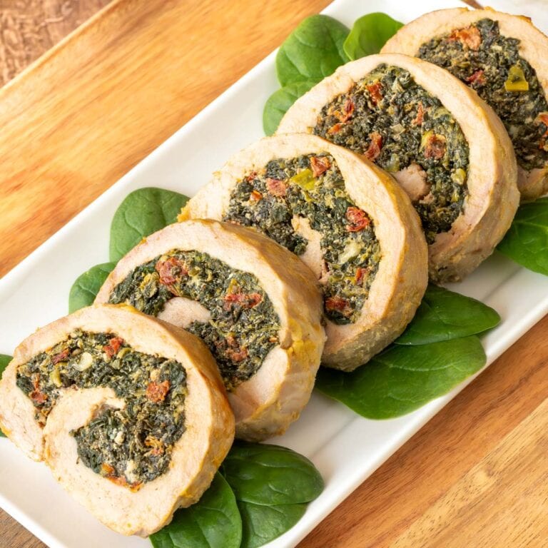 Five slices of stuffed pork tenderloin on a white platter, garnished with baby spinach on the side