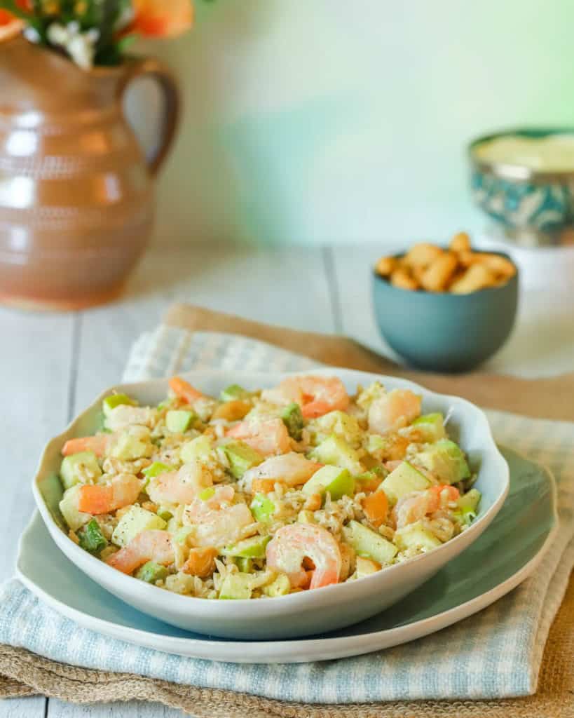 Chopped Shrimp Salad with Rice and Avocado mounded on top of a blue plate, with cashews and a pitcher if flowers in the background