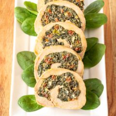 Five slices of stuffed pork tenderloin on a white platter, garnished with baby spinach on the side